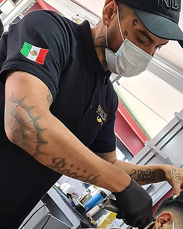 Barbero con gorra, cubrebocas y brazos tatuados trabajando con la máquina