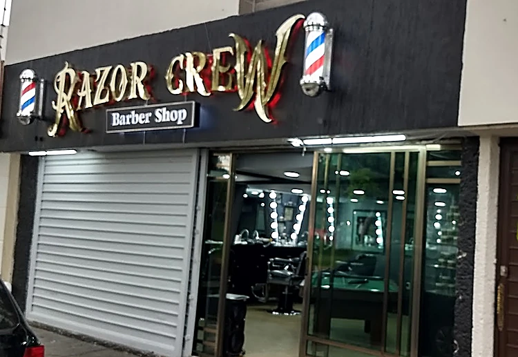 Fachada de Razor Crew: letrero negro con letras doradas, poste de barbero y, tras el vidrio, espejos con focos, sillas y la mesa de billar